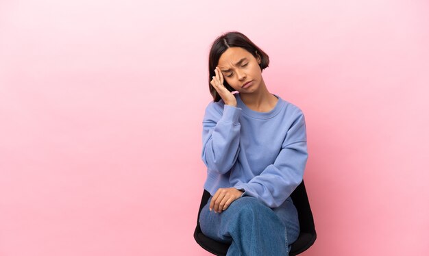 Joven mujer de raza mixta sentada en una silla aislada sobre fondo rosa con dolor de cabeza