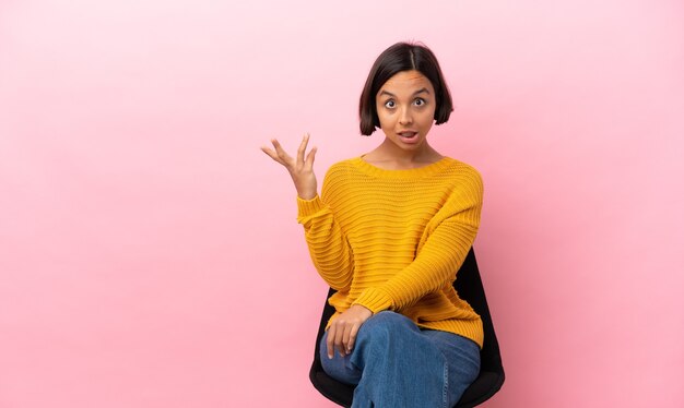 Joven mujer de raza mixta sentada en una silla aislada sobre fondo rosa haciendo gesto de dudas