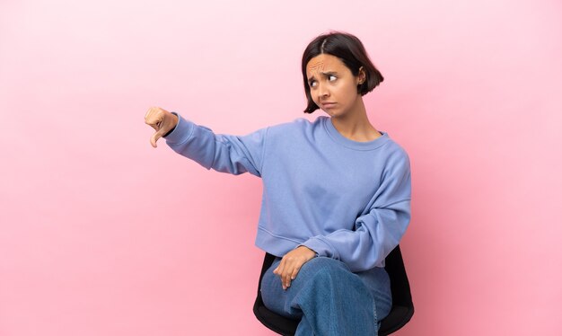 Joven mujer de raza mixta sentada en una silla aislada sobre fondo rosa mostrando el pulgar hacia abajo con expresión negativa