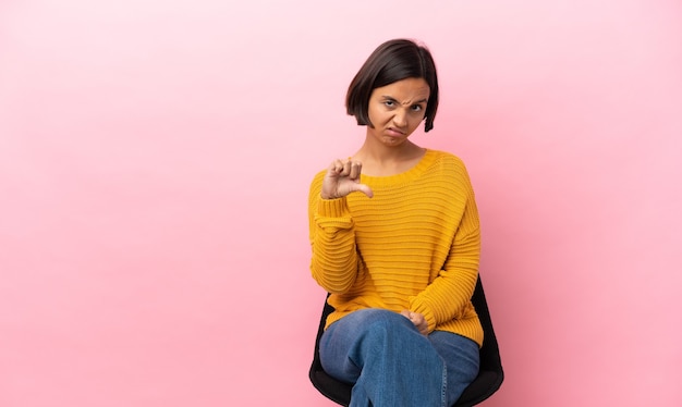 Joven mujer de raza mixta sentada en una silla aislada sobre fondo rosa mostrando el pulgar hacia abajo con expresión negativa