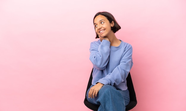 Joven mujer de raza mixta sentada en una silla aislada sobre fondo rosa pensando en una idea