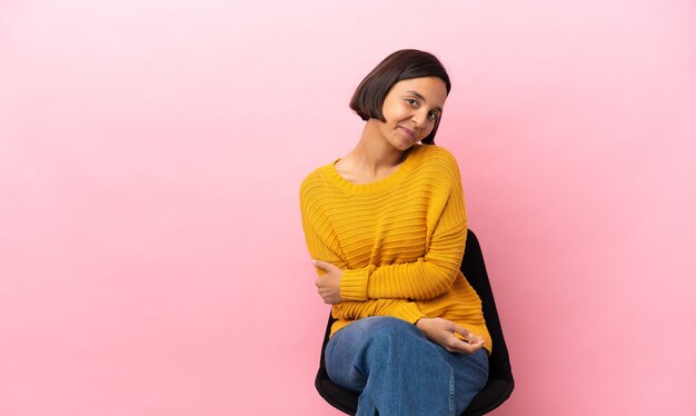 Joven mujer de raza mixta sentada en una silla aislada sobre fondo rosa riendo