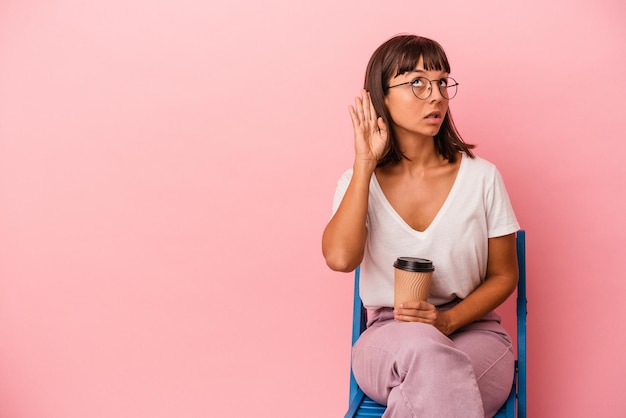 Foto joven mujer de raza mixta sentada en una silla sosteniendo un café aislado en un fondo rosado tratando de escuchar un chisme