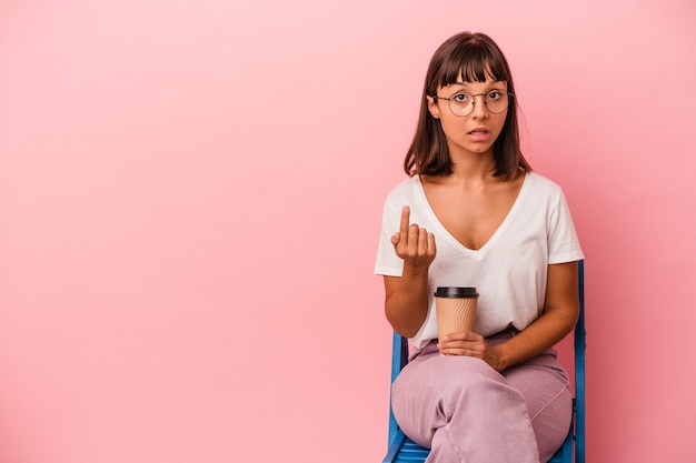 Joven mujer de raza mixta sentada en una silla sosteniendo un café aislado sobre fondo rosa apuntando con el dedo como si invitara a acercarse.