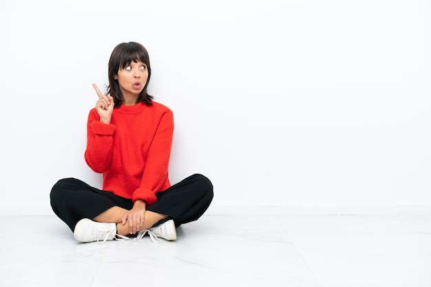 Joven mujer de raza mixta sentada en el suelo aislada de fondo blanco pensando en una idea apuntando con el dedo hacia arriba