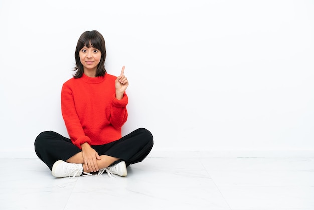 Joven mujer de raza mixta sentada en el suelo aislada de fondo blanco señalando con el dedo índice una gran idea