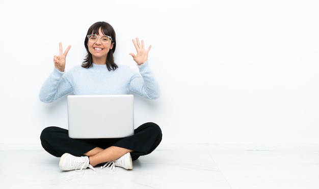 Joven mujer de raza mixta sentada en el suelo con una laptop aislada de fondo blanco contando siete con los dedos