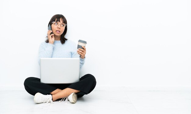 Joven mujer de raza mixta sentada en el suelo con una laptop aislada de fondo blanco sosteniendo café para llevar y un móvil