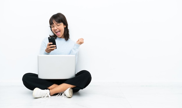 Joven mujer de raza mixta sentada en el suelo con una laptop aislada de fondo blanco con el teléfono en posición de victoria
