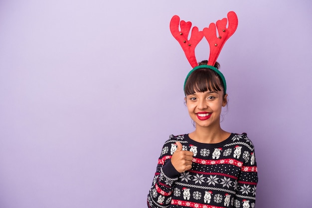 Joven mujer de raza mixta con sombrero de reno celebrando la Navidad aislado sobre fondo púrpura sonriendo y levantando el pulgar hacia arriba