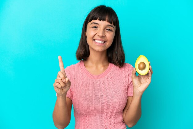 Joven mujer de raza mixta sosteniendo un aguacate aislado de fondo azul mostrando y levantando un dedo