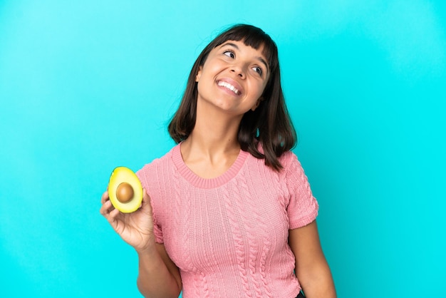 Joven mujer de raza mixta sosteniendo un aguacate aislado de fondo azul pensando en una idea mientras mira hacia arriba