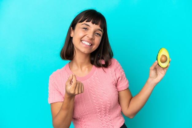 Joven mujer de raza mixta sosteniendo un aguacate aislado sobre fondo azul haciendo gesto de dinero