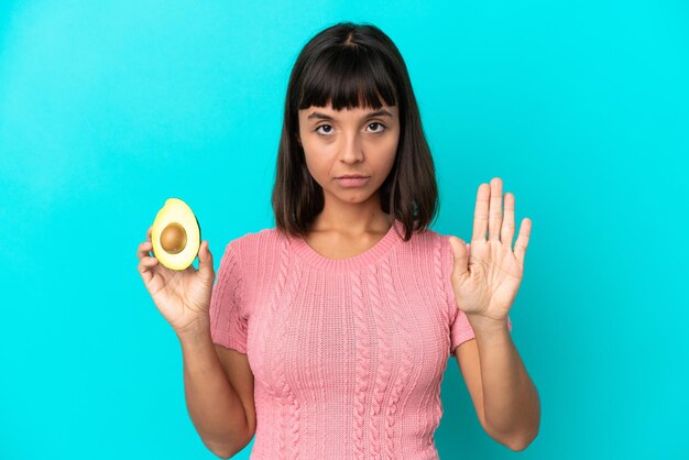 Joven mujer de raza mixta sosteniendo un aguacate aislado sobre fondo azul haciendo gesto de parada