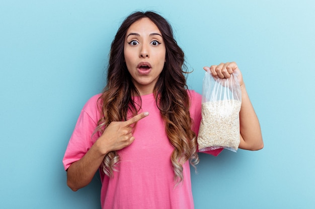 Joven mujer de raza mixta sosteniendo avena aislado sobre fondo azul apuntando hacia el lado