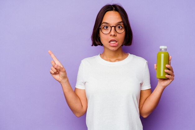 Joven mujer de raza mixta sosteniendo un batido saludable aislado sobre fondo púrpura apuntando hacia el lado