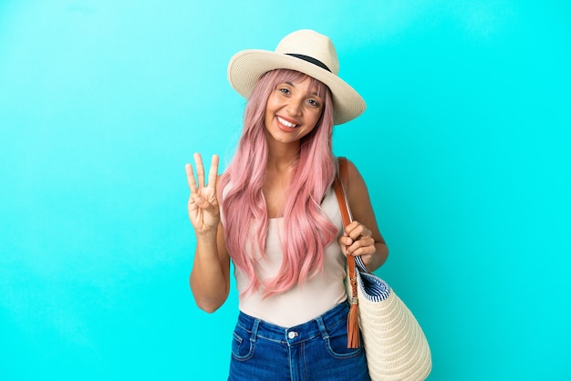 Foto joven mujer de raza mixta sosteniendo una bolsa de playa con pamela aislado sobre fondo azul feliz y contando tres con los dedos