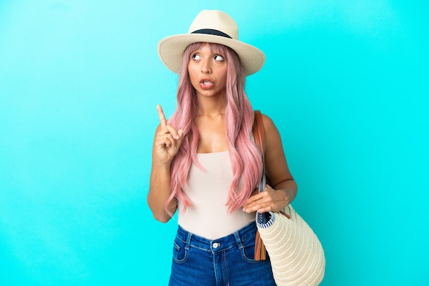 Joven mujer de raza mixta sosteniendo una bolsa de playa con pamela aislado sobre fondo azul pensando en una idea apuntando con el dedo hacia arriba