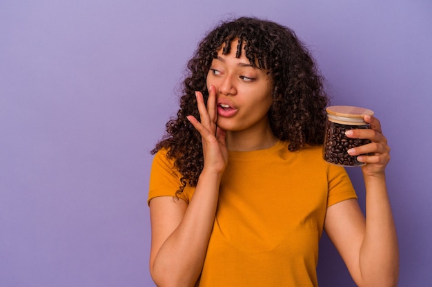 Joven mujer de raza mixta sosteniendo una botella de granos de café aislada sobre fondo púrpura está diciendo una noticia secreta de frenado en caliente y mirando a un lado