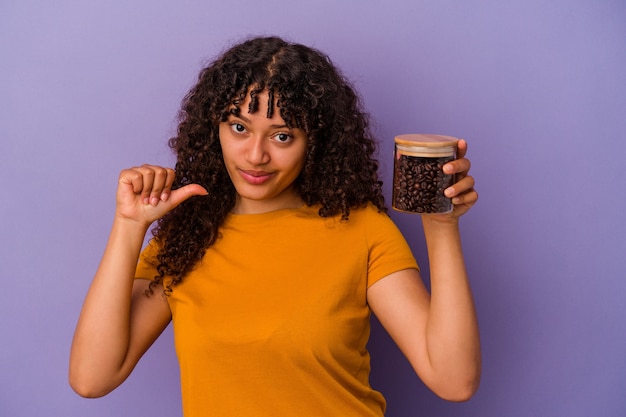 Joven mujer de raza mixta sosteniendo una botella de granos de café aislada sobre fondo púrpura se siente orgullosa y segura de sí misma, ejemplo a seguir.