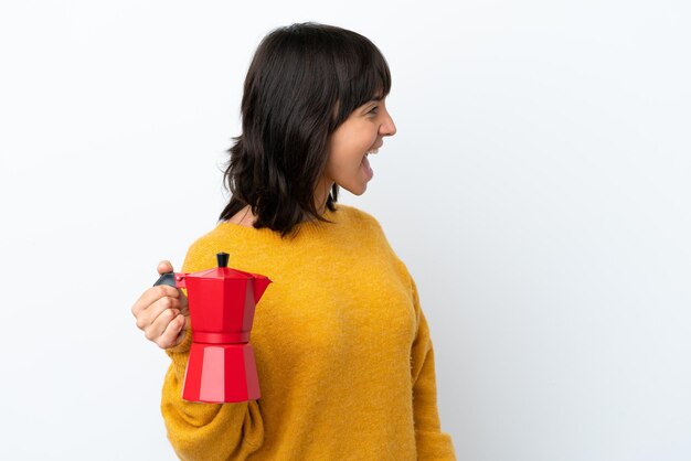 Joven mujer de raza mixta sosteniendo una cafetera aislada de fondo blanco riéndose en posición lateral