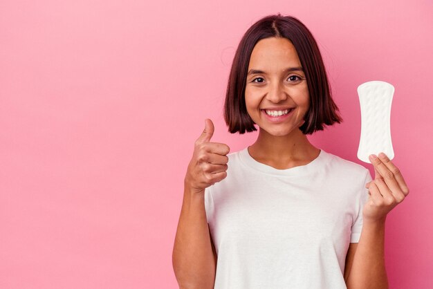 Joven mujer de raza mixta sosteniendo una compresa aislada sobre fondo rosa sonriendo y levantando el pulgar hacia arriba