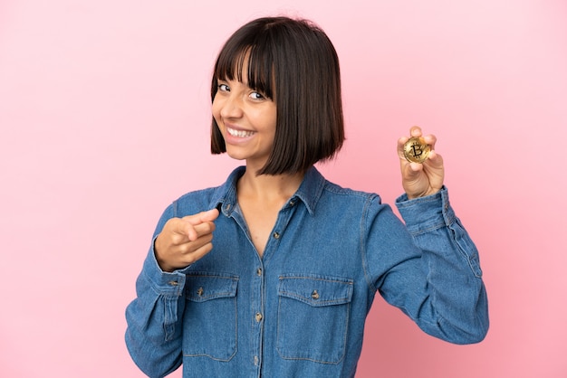 Joven mujer de raza mixta sosteniendo un fondo aislado Bitcoin apuntando hacia el frente con expresión feliz