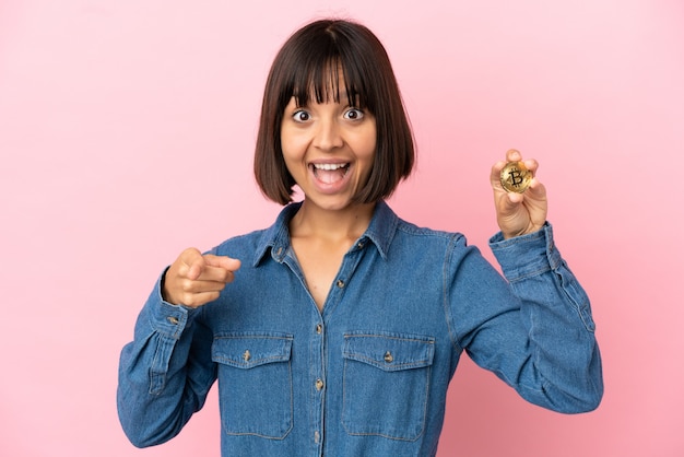 Joven mujer de raza mixta sosteniendo un fondo aislado de Bitcoin sorprendido y apuntando hacia el frente