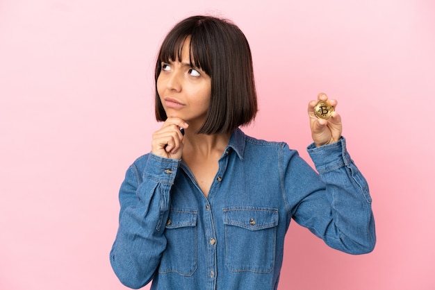 Joven mujer de raza mixta sosteniendo un fondo aislado de Bitcoin teniendo dudas