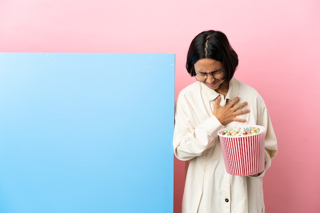 Joven mujer de raza mixta sosteniendo palomitas de maíz con una gran pancarta sobre antecedentes aislados con dolor en el corazón