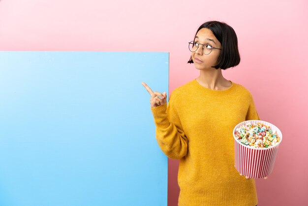 Joven mujer de raza mixta sosteniendo palomitas de maíz con una gran pancarta sobre fondo aislado apuntando hacia una gran idea