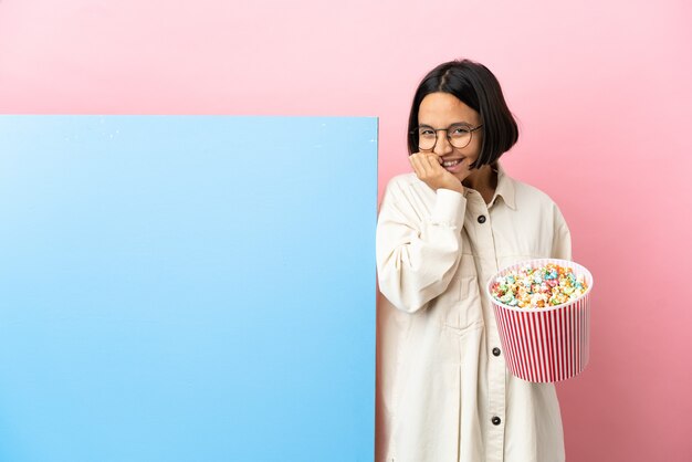 Joven mujer de raza mixta sosteniendo palomitas de maíz con una gran pancarta sobre fondo aislado feliz y sonriente cubriendo la boca con las manos