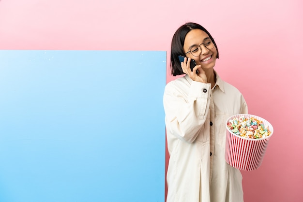 Joven mujer de raza mixta sosteniendo palomitas de maíz con una gran pancarta sobre fondo aislado manteniendo una conversación con el teléfono móvil