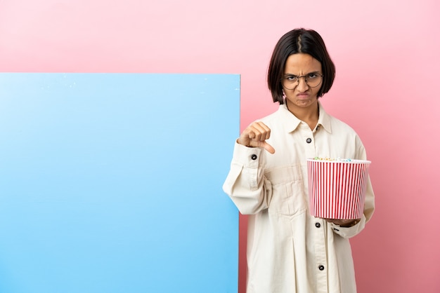 Foto joven mujer de raza mixta sosteniendo palomitas de maíz con una gran pancarta sobre fondo aislado mostrando el pulgar hacia abajo con expresión negativa