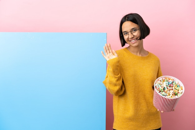 Joven mujer de raza mixta sosteniendo palomitas de maíz con una gran pancarta sobre fondo aislado saludando con la mano con expresión feliz