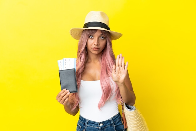 Joven mujer de raza mixta sosteniendo pasaporte y bolsa de playa aislada sobre fondo amarillo haciendo gesto de parada