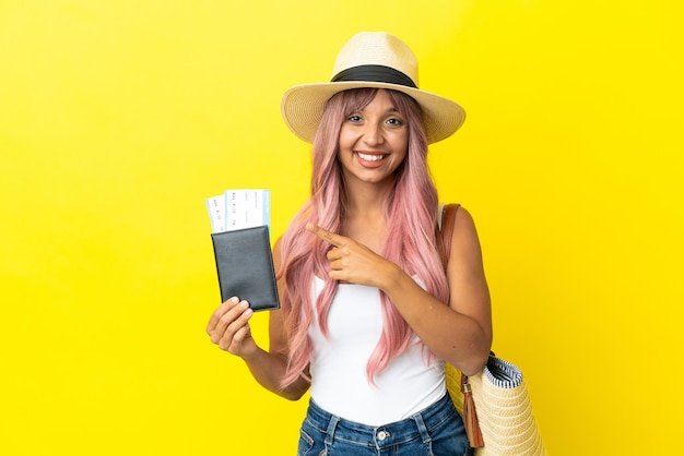 Joven mujer de raza mixta sosteniendo pasaporte y bolsa de playa aislado sobre fondo amarillo apuntando hacia el lado para presentar un producto