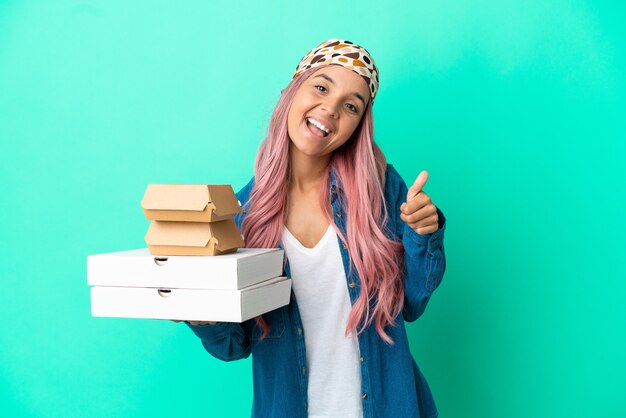 Joven mujer de raza mixta sosteniendo pizzas y hamburguesas aisladas sobre fondo verde con los pulgares hacia arriba porque algo bueno ha sucedido