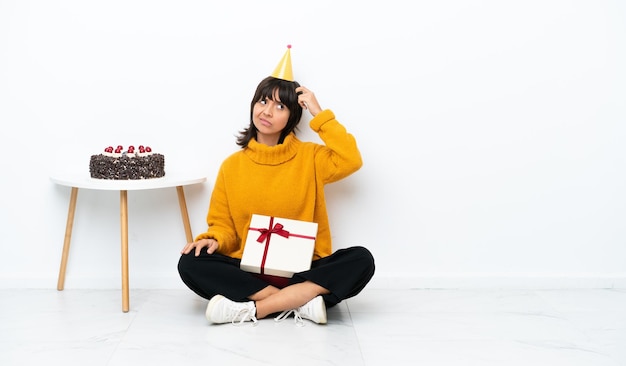 Joven mujer de raza mixta sosteniendo un regalo sentada en el suelo aislada de fondo blanco con dudas y con una expresión facial confusa