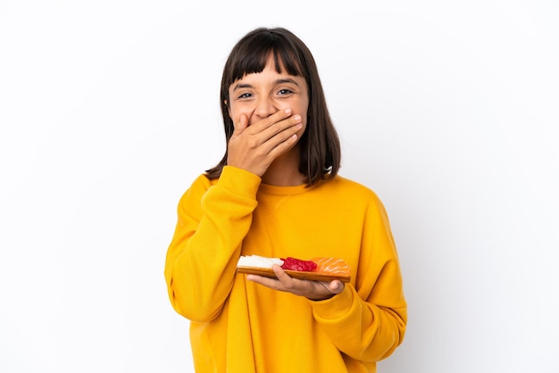 Joven mujer de raza mixta sosteniendo sashimi aislado sobre fondo blanco feliz y sonriente cubriendo la boca con la mano