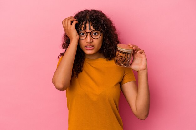 Joven mujer de raza mixta sosteniendo un tarro de almendras aislado sobre fondo rosa conmocionado, ella ha recordado una reunión importante.