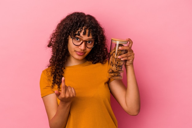 Joven mujer de raza mixta sosteniendo un tarro de galletas aislado sobre fondo rosa apuntando con el dedo hacia usted como si invitara a acercarse.