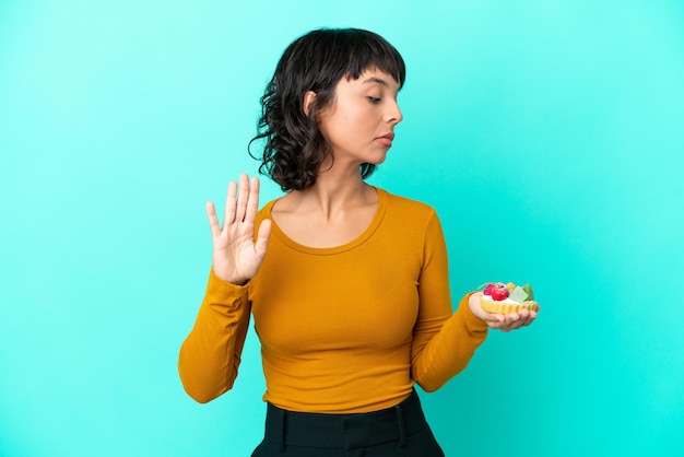 Foto joven mujer de raza mixta sosteniendo una tartaleta aislada de fondo azul haciendo un gesto de parada y decepcionada