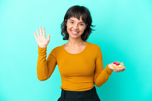 Joven mujer de raza mixta sosteniendo una tartaleta aislada de fondo azul saludando con la mano con expresión feliz