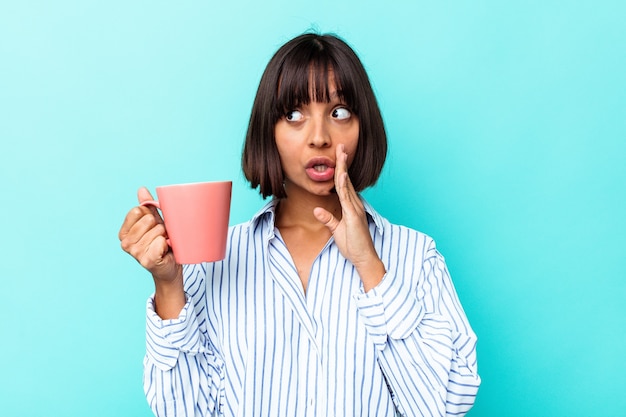 Joven mujer de raza mixta sosteniendo una taza rosa aislada sobre fondo azul está diciendo una noticia secreta de frenado en caliente y mirando a un lado