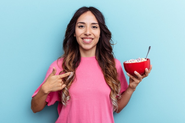 Joven mujer de raza mixta sosteniendo un tazón de cereales aislado sobre fondo azul persona apuntando con la mano a un espacio de copia de camisa, orgulloso y seguro