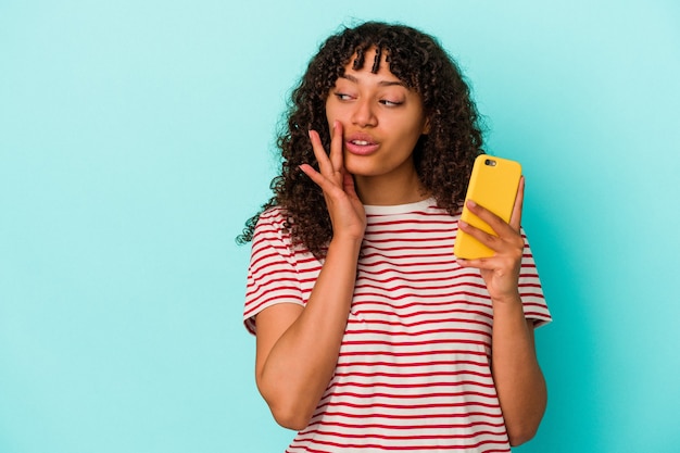 Joven mujer de raza mixta sosteniendo un teléfono móvil aislado sobre fondo azul está diciendo una noticia secreta de frenado en caliente y mirando a un lado