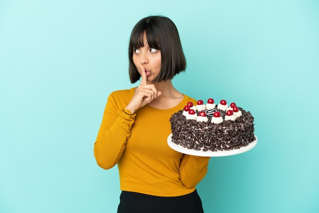 Joven mujer de raza mixta sosteniendo la torta de cumpleaños mostrando un gesto de silencio poniendo el dedo en la boca