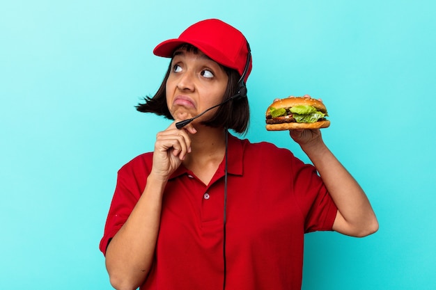 Joven mujer de raza mixta trabajador de restaurante de comida rápida sosteniendo una hamburguesa aislado sobre fondo azul.