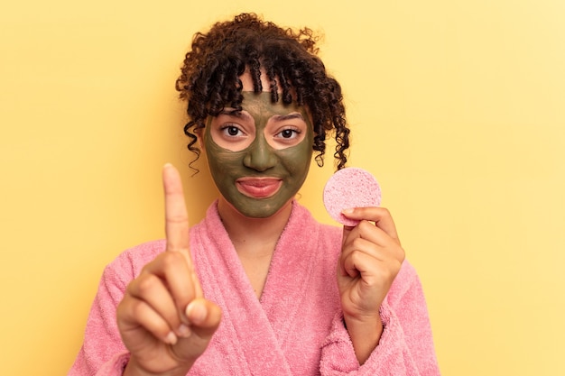 Joven mujer de raza mixta vistiendo una bata de baño sosteniendo una esponja desmaquillante aislada sobre fondo amarillo que muestra el número uno con el dedo.